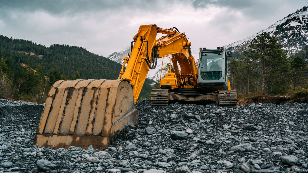 Excavator vehicle digging into rocky material in an area surrounded by mountains and trees