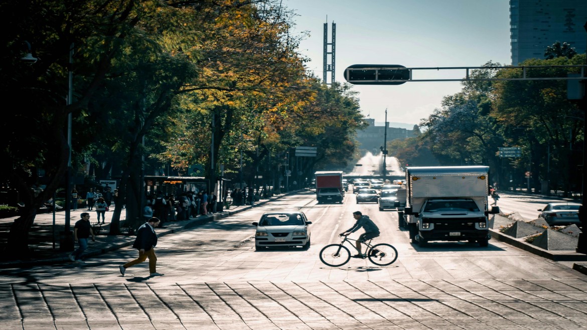 City view of a bicyclist crossing a street in front of a car and truck with trees on the left
